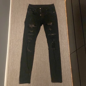 Svarta skinny jeans med slitningar och paljettdetaljer - Säljer ett par svarta skum fit jeans med flera slitningar framtill som är dekorerade med svarta paljetter. Jeansen har normal passform i midjan och stängs med dragkedja och knappar. Perfekta för dig som vill ha en edgy stil.
