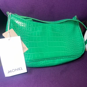 Grön axelväska från Monki (oanvänd) - Helt oanvänd snygg grön axelväska från Monki med prislapp fortfarande på. Väskan har en kedja och är i hekompakt form som passar perfekt till att bära det viktigaste. En trendig accessoar som sticker ut i mängden.