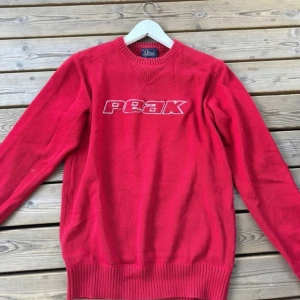 Röd stickad tröja från Peak - Röd stickad tröja från Peak med vit broderad logga på bröstet. Tröjan har rund halsringning och ribbade muddar vid ärmslut och nederkant. Perfekt för kyligare dagar.