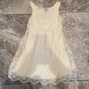 Vit spetsblus med knappar - Söt vit blus i spets med ärmlös design och dekorativa knappar framtill. Klänningen har rund halsringning och vacker spetskant nertill. Perfekt för somriga dagar eller speciella tillfällen.