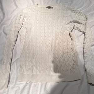 Vit kabelstickad tröja från Basic - Säljer en vit kabelstickad tröja från Basic i ekologisk bomull. Tröjan har rund halsringning, långa ärmar och ribbade muddar vid ärmslut och nederkant. Perfekt för lager-på-lager och enkel att matcha med olika outfits.