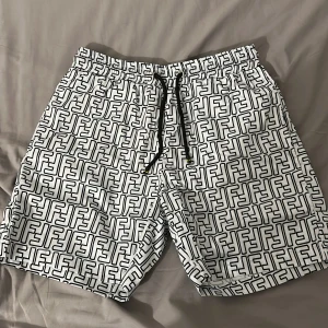Svarta och vita shorts från Fendi med FF-mönster - Snygga shorts från Fendi i vitt med svart FF-logomönster över hela tyget. De har elastisk midja med svart snörning och innerbyxa i mesh. Perfekta för sommaren och har en modern, loose passform. Det är Storlek S men den är felprintad så det står L