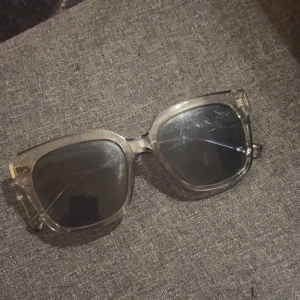 Grå transparenta solglasögon med bred båge - Stilrena solglasögon med transparent grå båge och svarta glas. Bågarna är breda och har en modern, fyrkantig form. Perfekta för att ge en cool touch till din outfit.