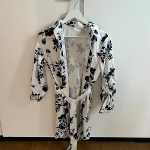 Vit och marinblå blommig omlottklänning - Säljer en vit omlottklänning med marinblå och svarta blommor. Klänningen har trekvartsärm, krage och knytband i midjan. Perfekt för en fräsch och stilren look med fina detaljer. Finns ingen lapp så vet ej märket elelr storlek men skulle gissa på storlek S💞