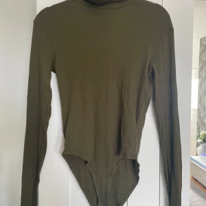 Olivgrön body - Säljer en olivgrön body med lång ärm. Bodyn är figurnära och har en hög hals, perfekt att styla med jeans eller kjol. Materialet är mjukt och stretchigt för en bekväm passform.