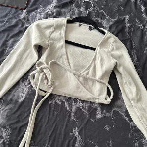 Beige omlottopp med knytband i färgen ljusgrön ish  - Trendig beige omlottopp med långa ärmar och knytband i midjan. Toppen är croppad och har en djup v-ringning. Perfekt att styla över ett linne eller topp.