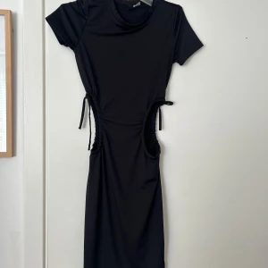 Svart kortärmad klänning med snörning i midjan - Säljer en svart klänning med korta ärmar och rund halsringning. Klänningen har snygga snörningsdetaljer i midjan som ger en cool och modern look. Perfekt för dig som gillar en stilren men trendig outfit.