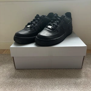 Svarta Nike Air Force 1 sneakers - Säljer ett par helsvarta Nike Air Force 1 sneakers. Klassisk modell med snörning, rund tå och platt sula. Skorna är i skinn och har en stilren look som passar till det mesta. Perfekta för dig som gillar streetwear och vill ha en ikonisk sneaker i garderoben.