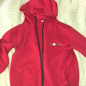 Röd hoodie från Boss - Röd hoodie från Boss med dragkedja och huva. Jackan har två fickor framtill och diskret Boss-logga på bröstet. Perfekt för en avslappnad stil.