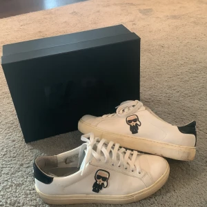Karl Lagerfeld vita sneakers  - Snygga vita sneakers från Karl Lagerfeld med svart häl och ikonisk Karl-figur på sidan. Skorna har snörning, rund tå och platt sula. Tillverkade i skinn och kommer med originalkartong. Perfekta för dig som gillar stilrena och coola detaljer.
