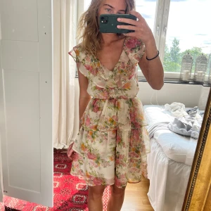Blommig volangklänning med v-ringning - Supersöt kort klänning med blommigt mönster i rosa, gult och grönt på en ljus botten. Klänningen har v-ringning, volanger och flera lager i kjolen som ger en flowy känsla. Ärmlös design med breda axelpartier och öppen rygg. Perfekt för sommarens alla tillfällen!