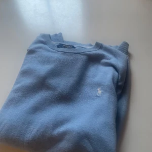 Ljusblå tröja från Polo Ralph Lauren - Säljer en ljusblå tröja från Polo Ralph Lauren med klassisk rund halsringning och broderad vit logga på bröstet. Tröjan är långärmad och har ribbade muddar vid ärmslut och nederkant. Perfekt för en clean och stilren look.
