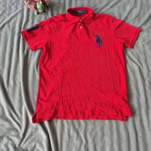 Röd pikétröja Polo Ralph Lauren M - Röd pikétröja från Polo Ralph Lauren med klassisk krage, två knappar och stor marinblå logga broderad på bröstet. På höger ärm finns en stor blå siffra 3. Tröjan är i bomull och har korta ärmar, perfekt för en sportig och stilren look.