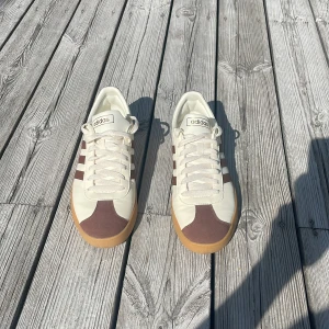 Adidas sneakers i beige och brunt - Snygga Adidas sneakers med beige ovandel, bruna detaljer och klassiska tre ränder på sidan. Skorna har gummisula, snörning och rund tå. Ovandelen är i en mix av syntet och mocka för en stilren look. Perfekta för dig som gillar retrovibbar och streetstyle.