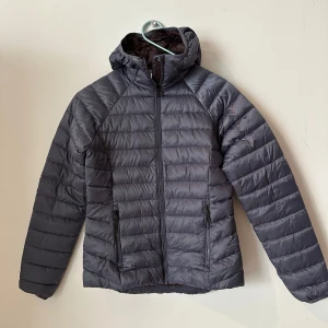 Grå/lila lättviksjacka från McKinley strl.34 - Snygg mörkgrå dunjacka från McKinley med huva och dragkedja framtill. Jackan är quiltad och har två praktiska fickor med dragkedja. Perfekt för kyliga dagar och enkel att matcha med olika outfits.