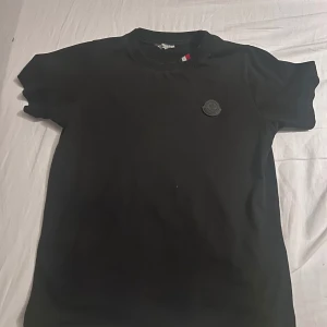 Svart Moncler t-shirt med logga - Svart t-shirt från Moncler med klassisk rund hals och korta ärmar. På bröstet sitter en svart Moncler-logga i gummi och i nacken finns en snygg detalj med röd, vit och blå rand. T-shirten är i mjuk bomull och har en clean, stilren look.