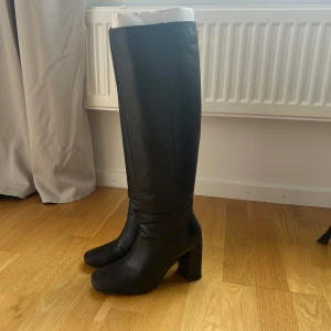 Svarta höga stövlar med klack - Snygga svarta stövlar i skinn med hög, blockig klack och rund tå. Stövlarna är knähöga och har en stilren, minimalistisk design som passar perfekt till både kjol och jeans. Använt två gånger, nypris 1499 i storlek 37 från nakd 