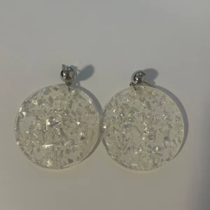 Stora glittriga örhängen i silver och vitt - Säljer ett par stora runda örhängen med glittrig effekt. De har en silvrig plupp upptill och en rund, transparent  platta med glittrande detaljer som reflekterar ljuset. Perfekt för dig som vill sticka ut och älskar statement-smycken.