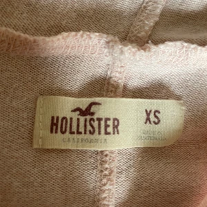 Ljusrosa hoodie från Hollister XS - Säljer en ljusrosa hoodie från Hollister i storlek XS. Tröjan har långa ärmar med två vita ränder på varje ärmslut, huva med snörning och en loose passform. Materialet är mjukt och tunt, perfekt för vår eller sommarkvällar. Superfin färg och chill stil!
