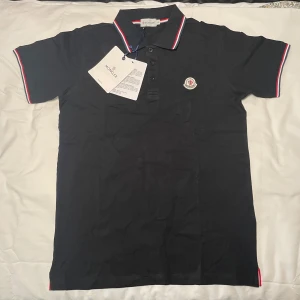 Moncler polotröja - Svart t-shirt från Moncler med klassisk pikékrage och knappslå. Kragen och ärmsluten har snygga ränder i rött, vitt och blått. Moncler-logga broderad på bröstet. Tillverkad i mjuk bomull, perfekt för en clean och sportig look. Finns två i storlek M och två i storlek L