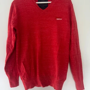 Röd stickad tröja från Superdry - Röd stickad tröja från Superdry med broderad logga och japanska tecken på bröstet. V-ringad modell med lång ärm och ribbade muddar. Perfekt för dig som gillar en enkel men snygg stil med lite edge.