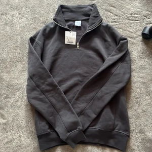 Mörkgrå half zip tröja från Zara - Snygg mörkgrå långärmad tröja från Zara med half zip och hög krage. Perfekt för dig som gillar en clean och enkel stil. Tröjan är gjord i mjuk bomull och har ribbade muddar vid ärmslut och nederkant. Storlek S. Helt oanvänd med lapp kvar.