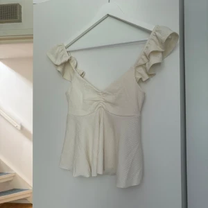 Beige blus med volangärmar - Supersöt beige blus med v-ringning och volangärmar. Toppen har en lätt rynkning vid bysten och ett flowy fall nedtill. Perfekt för dig som gillar romantiska detaljer och en feminin look.
