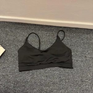 Svart ärmlös sportbh med smala band - Säljer en svart, enkel och stilren sportbh med smala axelband och v-ringad front. Toppen är croppad och har en twistad detalj baktill. Perfekt till träning