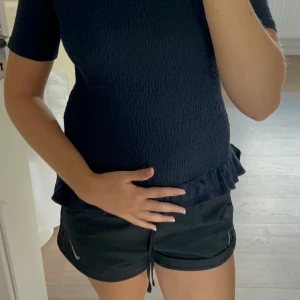 Mörkblå topp med volang från Gina Tricot - Supersöt mörkblå topp från Gina Tricot med korta ärmar och rund hals. Toppen har smockad struktur över hela kroppen och en liten volangkant längst ner. Perfekt att styla med jeans.