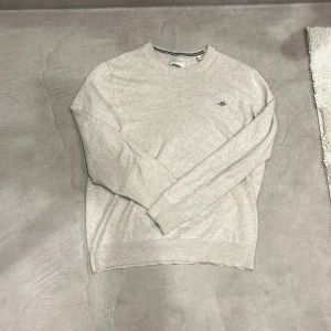 Beige stickad tröja från GANT - Snygg beige stickad tröja från GANT i storlek S. Tröjan har rund hals med diskret svart rand på insidan och GANTs broderade logga på bröstet. Perfekt för dig som gillar klassisk och clean stil. Långärmad och mjuk i materialet.