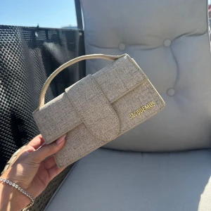 Beige handväska i linne - Säljer en stilren beige handväska  i linne med guldiga bokstäver på framsidan. Väskan har ett elegant, rektangulärt format och ett kort handtag. Perfekt för dig som gillar minimalistisk och trendig design. Band tillkommer 