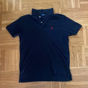 Mörkblå Polo Ralph Lauren t-shirt - Snygg mörkblå t-shirt från Polo Ralph Lauren med klassisk krage och två knappar framtill. Custom slim fit för en modern siluett. Ikonisk röd logobrodyr på bröstet. Perfekt för dig som gillar stilrena och tidlösa plagg.