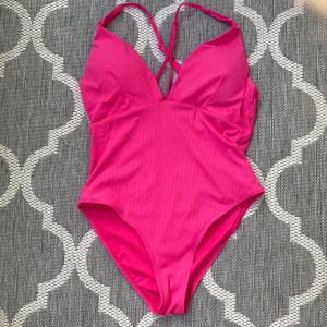 Rosa ribbad baddräkt med smala axelband - Snygg rosa baddräkt med ribbad struktur och smala, korsade axelband i ryggen. Djup v-ringning framtill och hög benskärning ger en modern look. Tillverkad i stretchigt material som sitter skönt på kroppen. Perfekt för strand eller poolhäng!