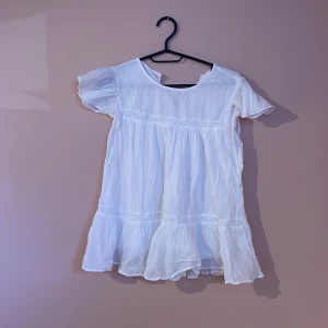 Vit kortklänning från Gina Tricot - Supersöt vit kortklänning från Gina Tricot med volangkant och lätt rynkad struktur. Klänningen har korta ärmar, rund hals framtill och v-ringad rygg. Tillverkad i mjukt bomullstyg som känns luftigt och fräscht. Perfekt för varma dagar och sommarhäng.