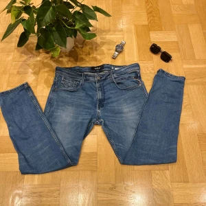 Replay Anbass blå jeans slim fit - Säljer ett par Replay Anbass jeans i klassisk blå tvätt med snygga slitningar. Modellen har slim fit och raka ben, fem fickor och detaljerade bakfickor med Replay-logga. Jeansen är tillverkade i stretchigt bomullsmaterial för skön passform.