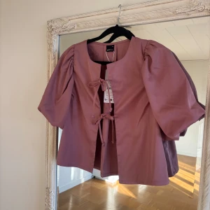 Blus med puffärm XS - Säljer en mauvefärgad overshirt från Gina Tricot i storlek XS. Jackan har puffiga ärmar, rund halsringning och knytning framtill. Perfekt för dig som gillar statement-ärmar och en feminin look. Materialet är mjukt och lätt, vilket gör den skön att bära.