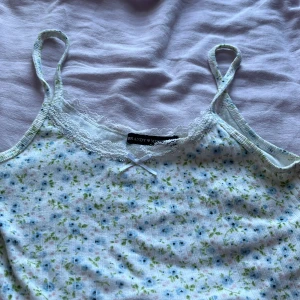 Blommig linnetopp från Brandy Melville - Supersöt linnetopp med blommigt mönster i blått, grönt och rosa på vit botten. Spaghettiband, spetskant vid urringningen och en liten rosett framtill. Ribbad bomullskvalitet och croppad passform – perfekt för varma dagar.