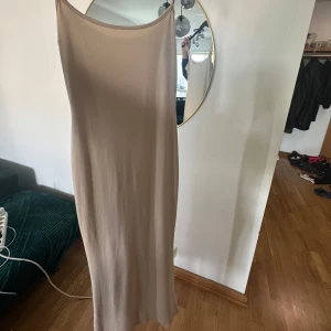 Beige långklänning med smala axelband - En stilren beige långklänning med smala axelband och rak passform. Klänningen har minimalistisk design och är perfekt för dig som gillar en enkel men elegant look. Materialet känns mjukt och följsamt mot kroppen.