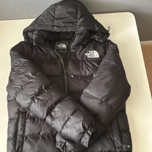 Svart pufferjacka från The North Face - Säljer en svart pufferjacka från The North Face i storlek L. Jackan har huva, dragkedja och två sidofickor. Klassisk logga på bröstet och ryggen. Perfekt för kalla dagar och riktigt snygg streetstil.