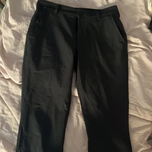 Svarta golfbyxor från Under Armour - Snygga svarta golfbyxor från Under Armour i klassisk rak modell. Byxorna har hällor för bälte, snedställda fickor fram och stängs med knapp och dragkedja. Perfekta för en clean och stilren look. Materialet är mjukt och följsamt.