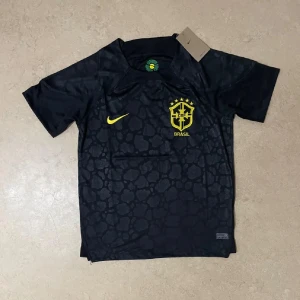 Svart Brasilien fotbollströja Nike - Snygg svart fotbollströja från Nike med Brasilien-emblem och gula detaljer. Tröjan har korta ärmar och ett diskret mönster över hela tyget. Perfekt för dig som vill sticka ut på planen eller visa ditt stöd för Brasilien.