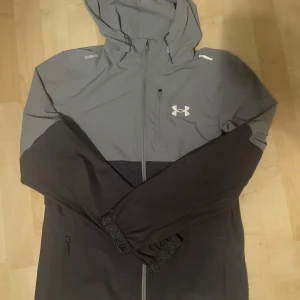 Under Armour grå och svart vindjacka - Snygg vindjacka från Under Armour i grått och svart med huva och dragkedja hela vägen. Jackan har reflekterande detaljer på axlarna och logga på bröstet. Perfekt för blåsiga dagar och sportiga outfits. Köpt från JD och har använt max 5gånger
