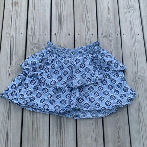 Blå mönstrad volangkjol Gina Tricot - Supersöt kort kjol från Gina Tricot i blått med vitt och mörkblått rutmönster. Kjolen har bred smock i midjan och dubbla volanger.