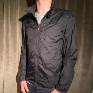 J.Lindeberg - Windbreaker  - Säljer en stilren svart vindjacka från J.Lindeberg. Jackan har en dragkedja framtill och är perfekt för höst och vår. Dragkedjan har gått av på mitten (se bild 5) men den går fortfarande att stänga och inget som stör användning. Vid minsta fråga eller fundering hör av er🤝