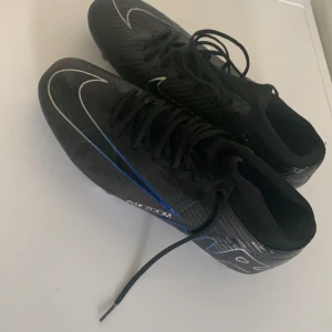 Svarta Nike Air Zoom fotbollsskor - Säljer ett par svarta Nike Air Zoom fotbollsskor med snygga vita och blå detaljer. Skorna har snörning och är designade för att ge bra grepp på planen. Perfekta för dig som vill ha stilrena och sportiga skor till matchen.