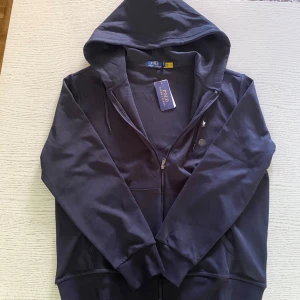 Mörkblå hoodie från Polo Ralph Lauren - Mörkblå hoodie från Polo Ralph Lauren med dragkedja och huva. Klassisk design med vit broderad logga på bröstet och snörning vid huvan. Tillverkad i mjuk bomull, perfekt för chill dagar. Fickor framtill och ribbade muddar för extra komfort.
