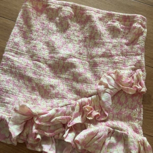 Rosa blommig kortkjol med volang - Supersöt kortkjol i rosa och vit med blommigt mönster och volangdetaljer längs nederkanten. Kjolen har en smockad struktur som ger en skön och stretchig passform. Perfekt för dig som gillar romantisk och lekfull stil.