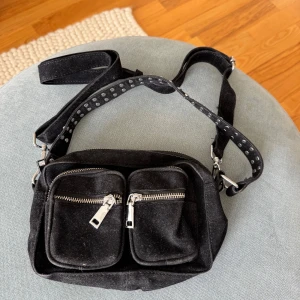 Svart axelväska med nitar och dragkedjor - Cool svart axelväska i mocka med två framfickor och silvriga dragkedjor. Axelremmen är dekorerad med nitar för en edgy vibe. Perfekt storlek för mobilen, plånboken och lite till. Snygg och praktisk för dig som gillar streetstyle.