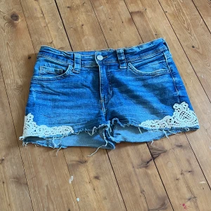 Blå jeansshorts med spetsdetaljer - Snygga blå jeansshorts med rå kant och vita spetsdetaljer på benen. Klassisk femficksmodell med knapp och dragkedja framtill. Perfekt för dig som gillar en chill och trendig stil med lite extra detaljer.