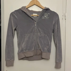Grå velour hoodie från Juicy Couture - Supermjuk grå hoodie från Juicy Couture i velour med glittrigt silvertryck fram och bak. Trycket har blivit slitet som ni ser på bilderna men frf super fin passar strl 134-140. Klassisk huva, dragkedja och två fickor framtill. Perfekt för en chill och trendig look.